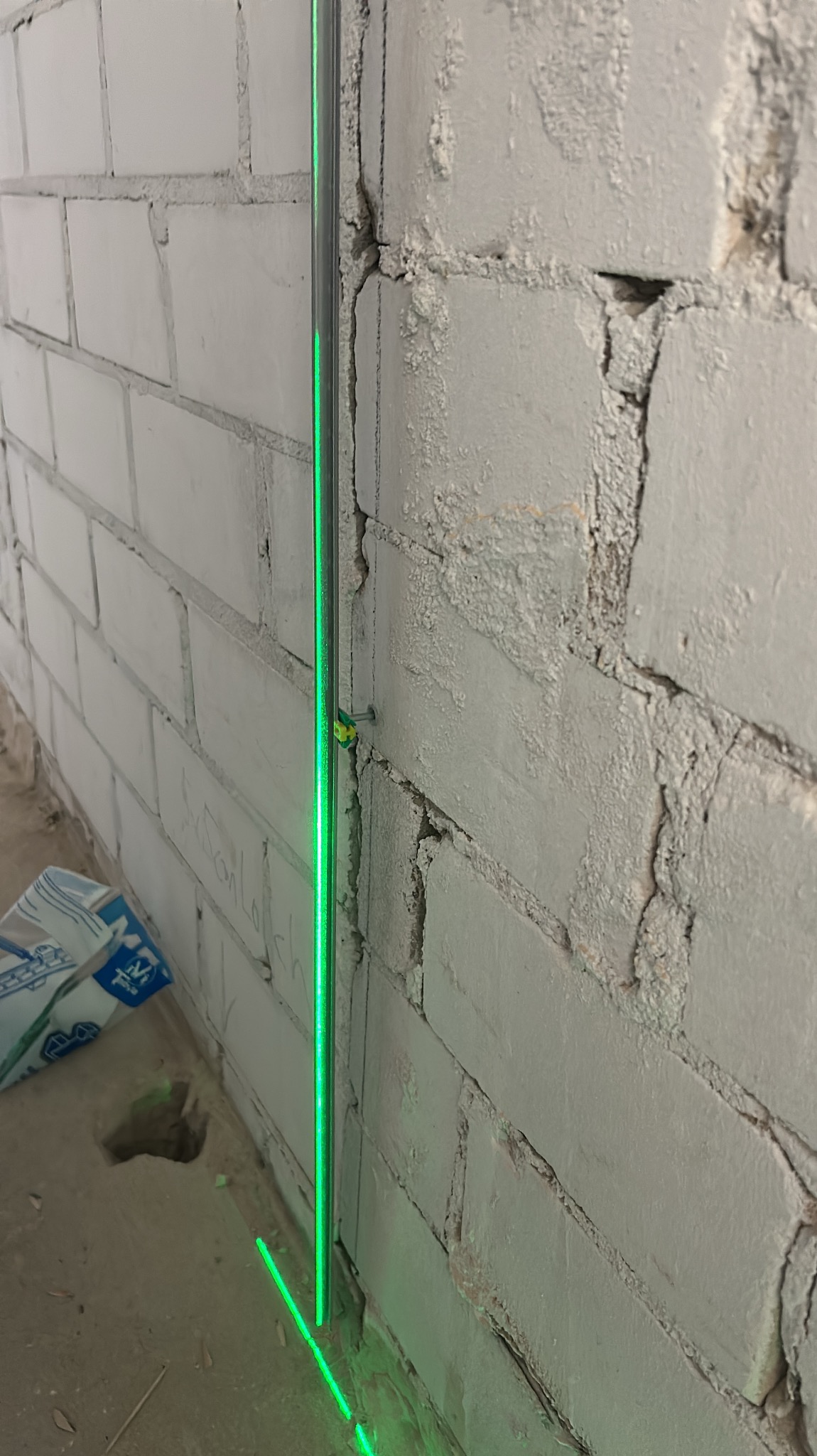 Ceglana ściana z widocznym pionem lasera, przymocowanym do ściany za pomocą gwoździa, wskazującym linię prostą; w tle widoczny worek z zaprawą.