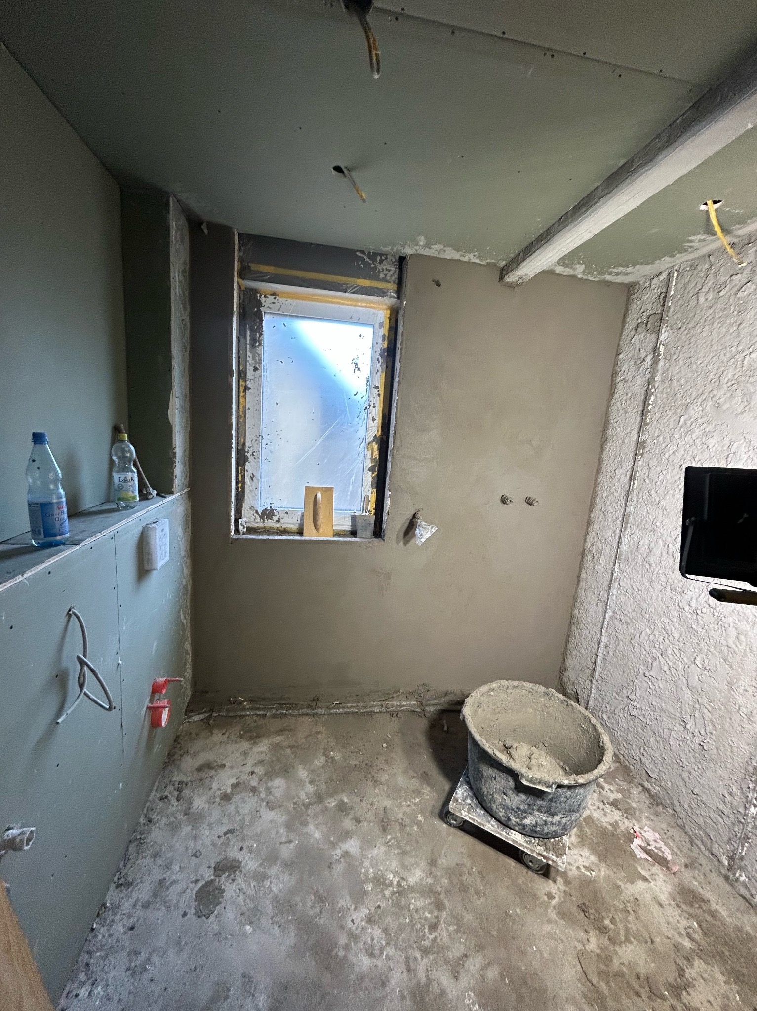 Pokój w trakcie prac wykończeniowych: ściany i sufit pokryte świeżym tynkiem, widoczne butelki z wodą na półce, wiadro z zaprawą na podłodze, okno zabezpieczone folią.