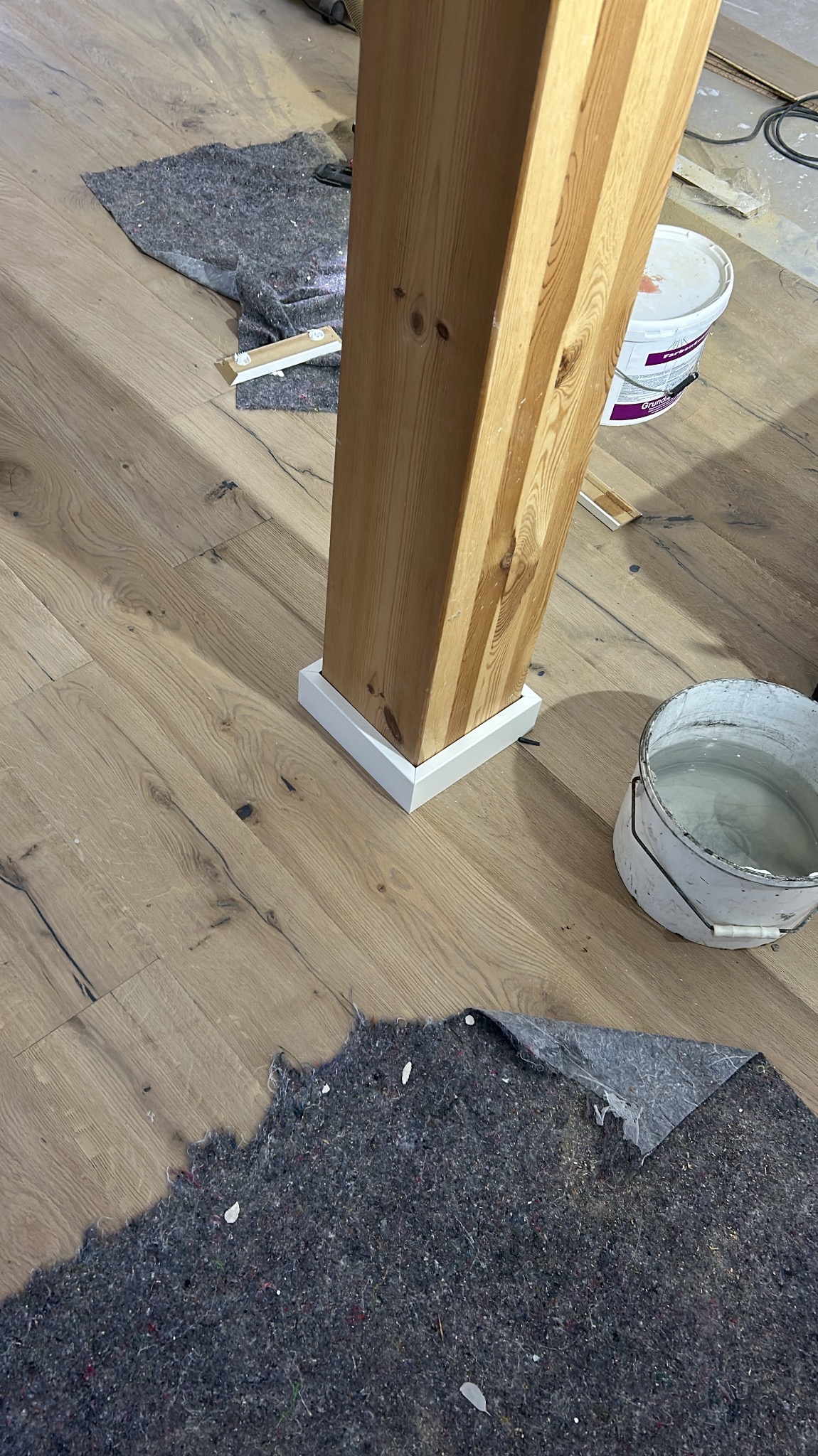 Drewniany słup z białą listwą przypodłogową na nowej drewnianej podłodze, zabezpieczony materiałem ochronnym i wiadrem z farbą w tle.