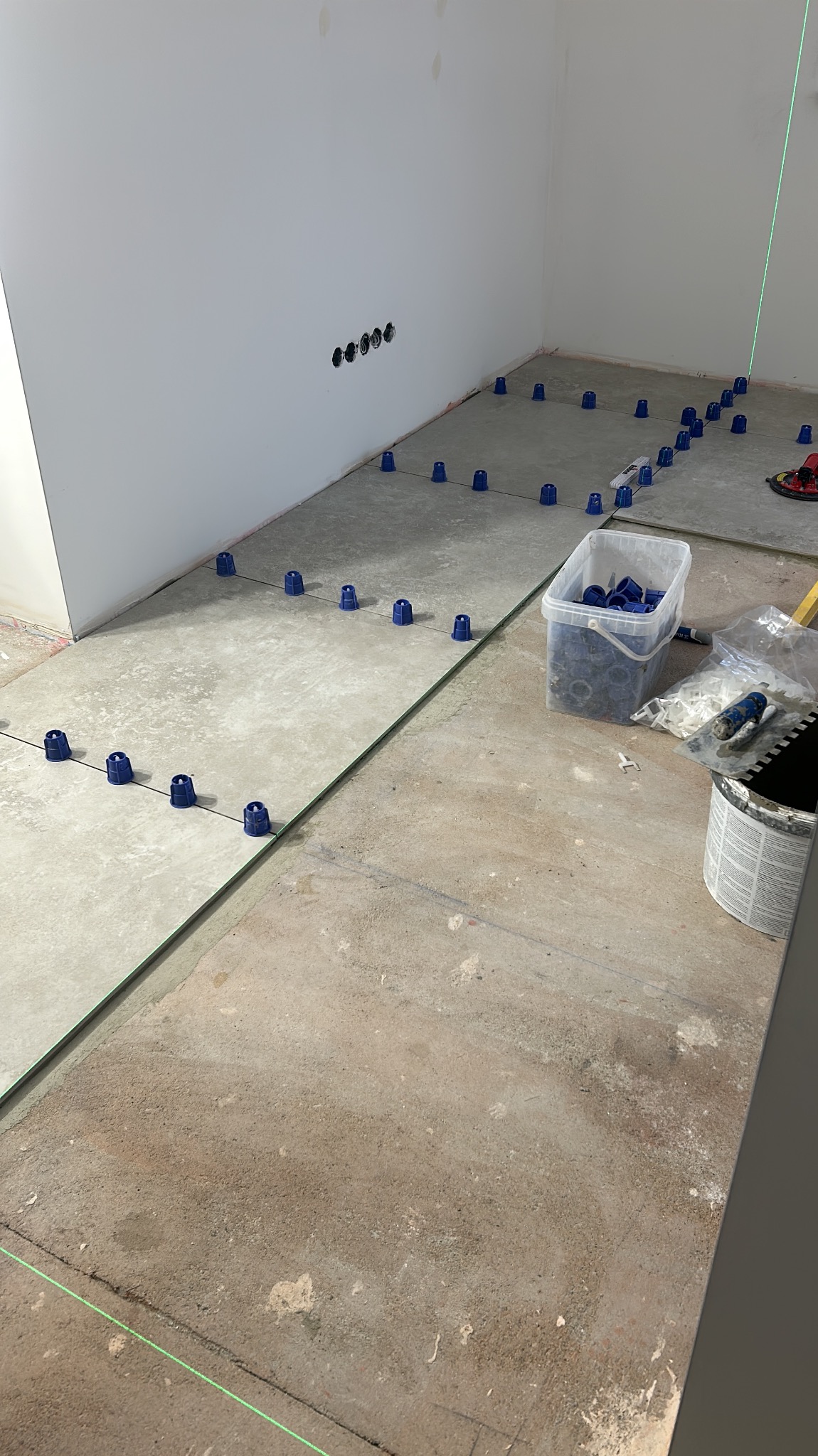 Układanie płytek podłogowych z użyciem systemu poziomowania, widoczne niebieskie klipsy, poziom laserowy, pojemnik z klipsami, paca do nakładania kleju i surowa podłoga.