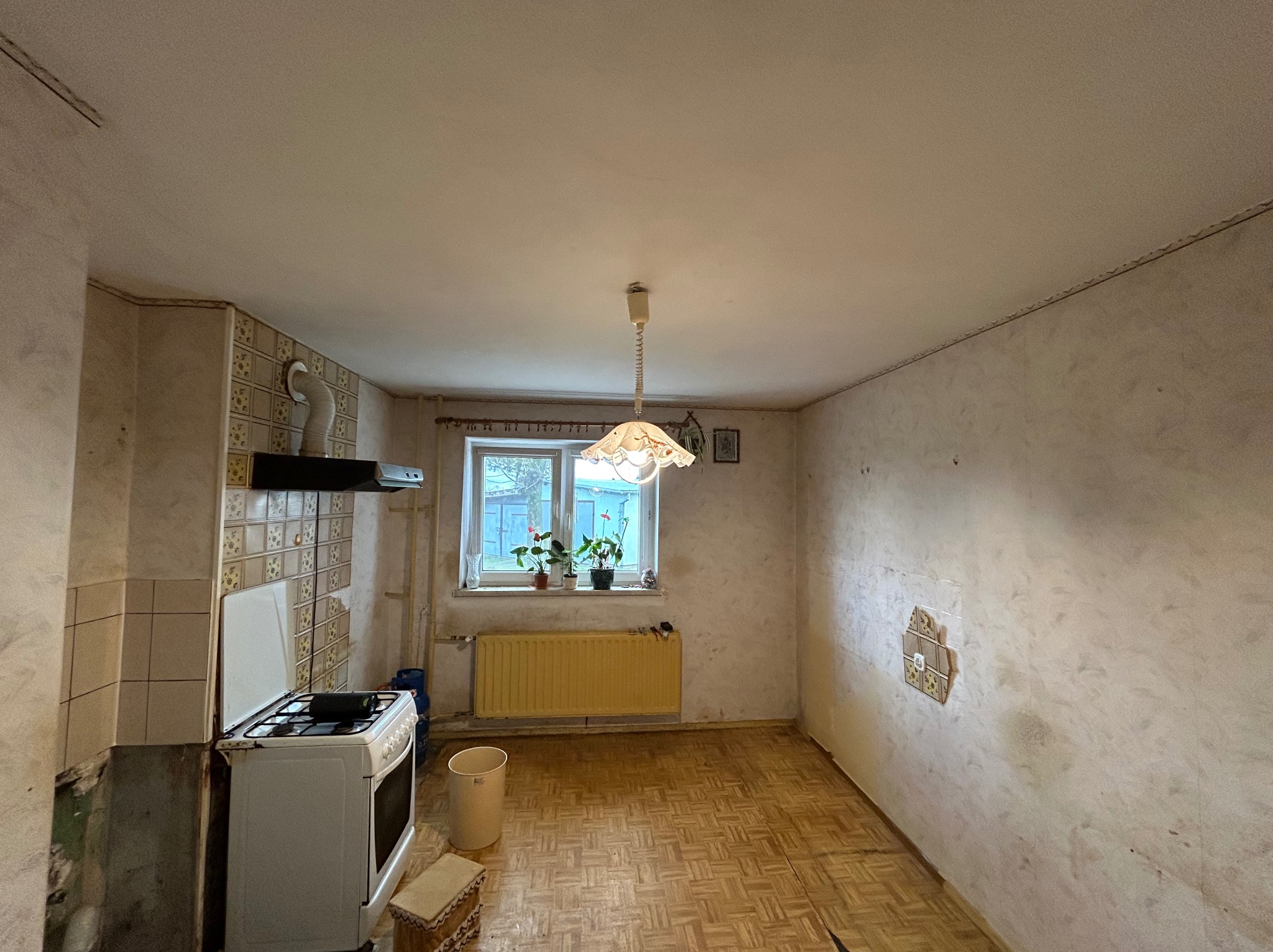 Wnętrze kuchni przed remontem: widoczne stare płytki, zniszczone ściany, żółty grzejnik pod oknem, kuchenka gazowa i parkiet w jodełkę.