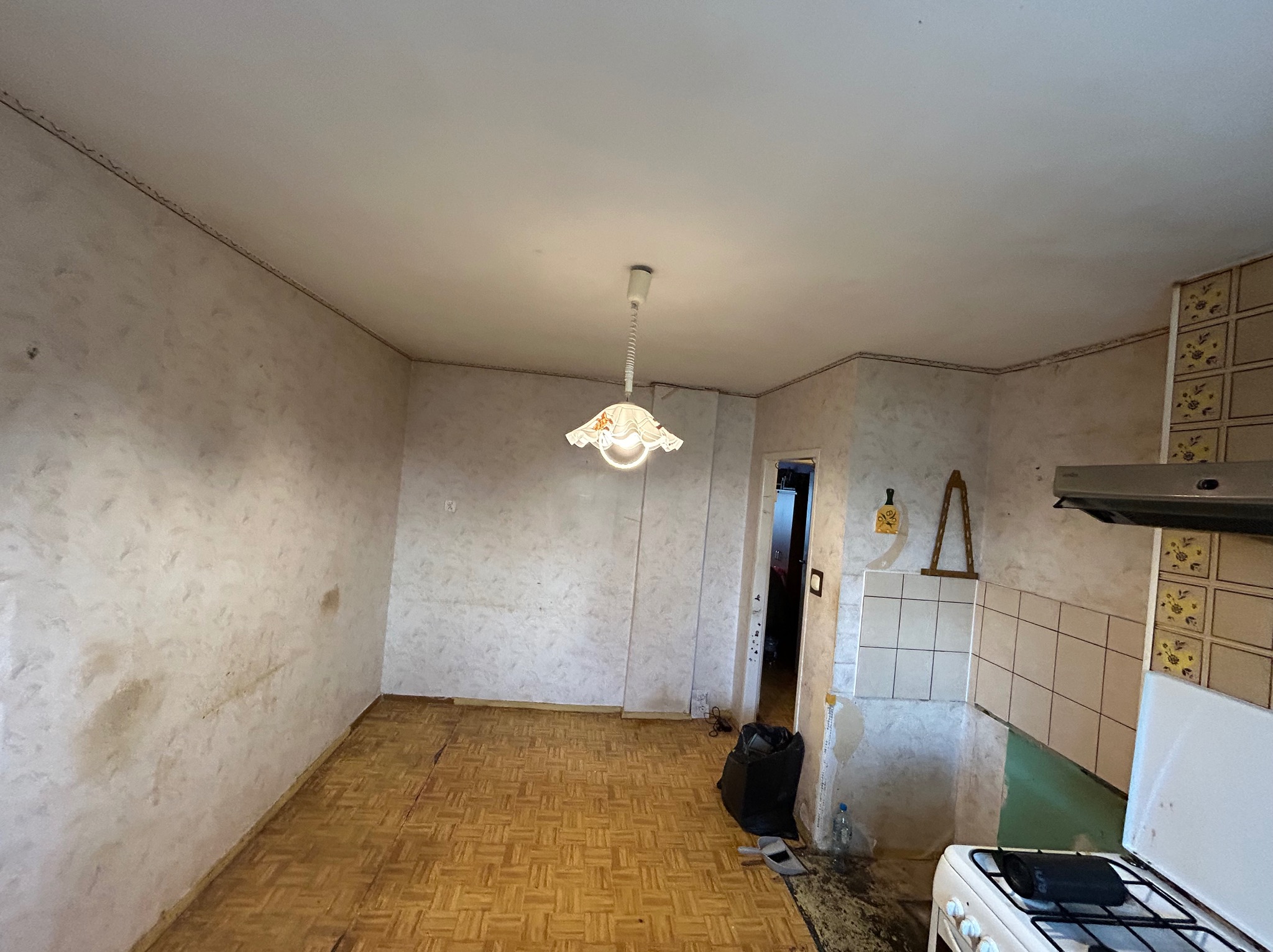Kuchnia w trakcie remontu: zerwane stare płytki, widoczne ślady po kleju na ścianach, parkiet w stanie wymagającym odnowienia, wisząca lampa z abażurem w kwiaty, śmieci w worku, stara kuchenka.