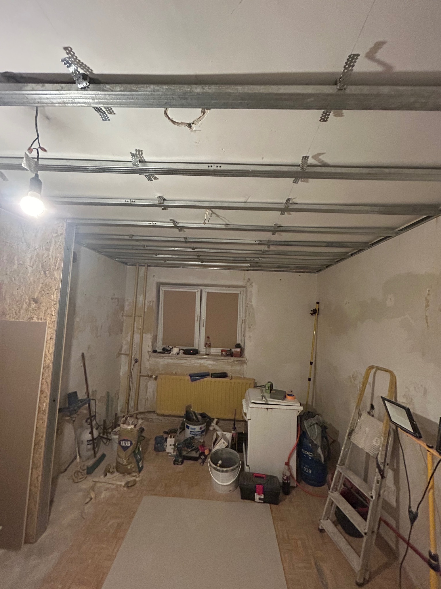 Montaż metalowego stelaża pod płytę gipsowo-kartonową na suficie w trakcie remontu. Widoczne profile, wieszaki, instalacja elektryczna, narzędzia i materiały budowlane w pomieszczeniu.