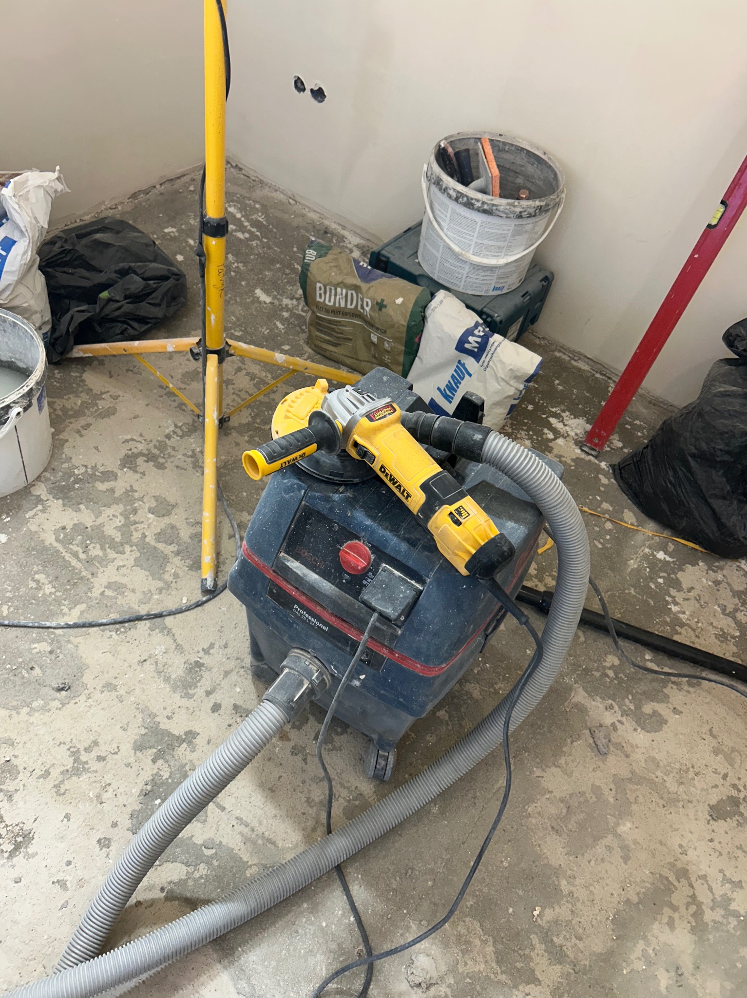 Żółta szlifierka DeWalt z podłączonym odkurzaczem przemysłowym na tle worków z zaprawą, poziomicy i lampy budowlanej w trakcie prac wykończeniowych.