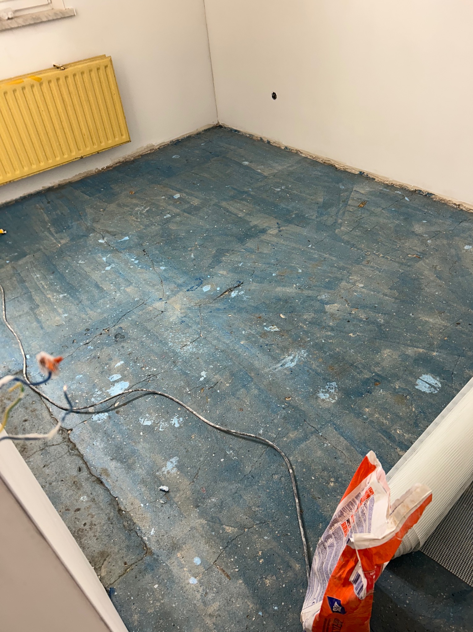 Przygotowanie podłoża: Niebieska, popękana wylewka w pokoju z żółtym grzejnikiem, widoczne kable i resztki materiałów budowlanych, sugerujące prace remontowe.