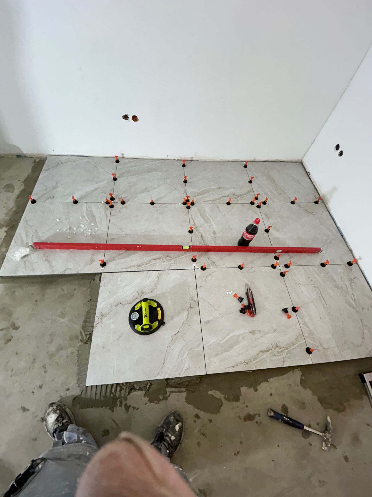 Układanie płytek podłogowych w trakcie prac: poziomica, przyssawka do płytek, butelka Coca-Coli, nożyk i młotek na podłodze z częściowo ułożonymi płytkami. Widoczne spoiwa i krzyżyki dystansowe.