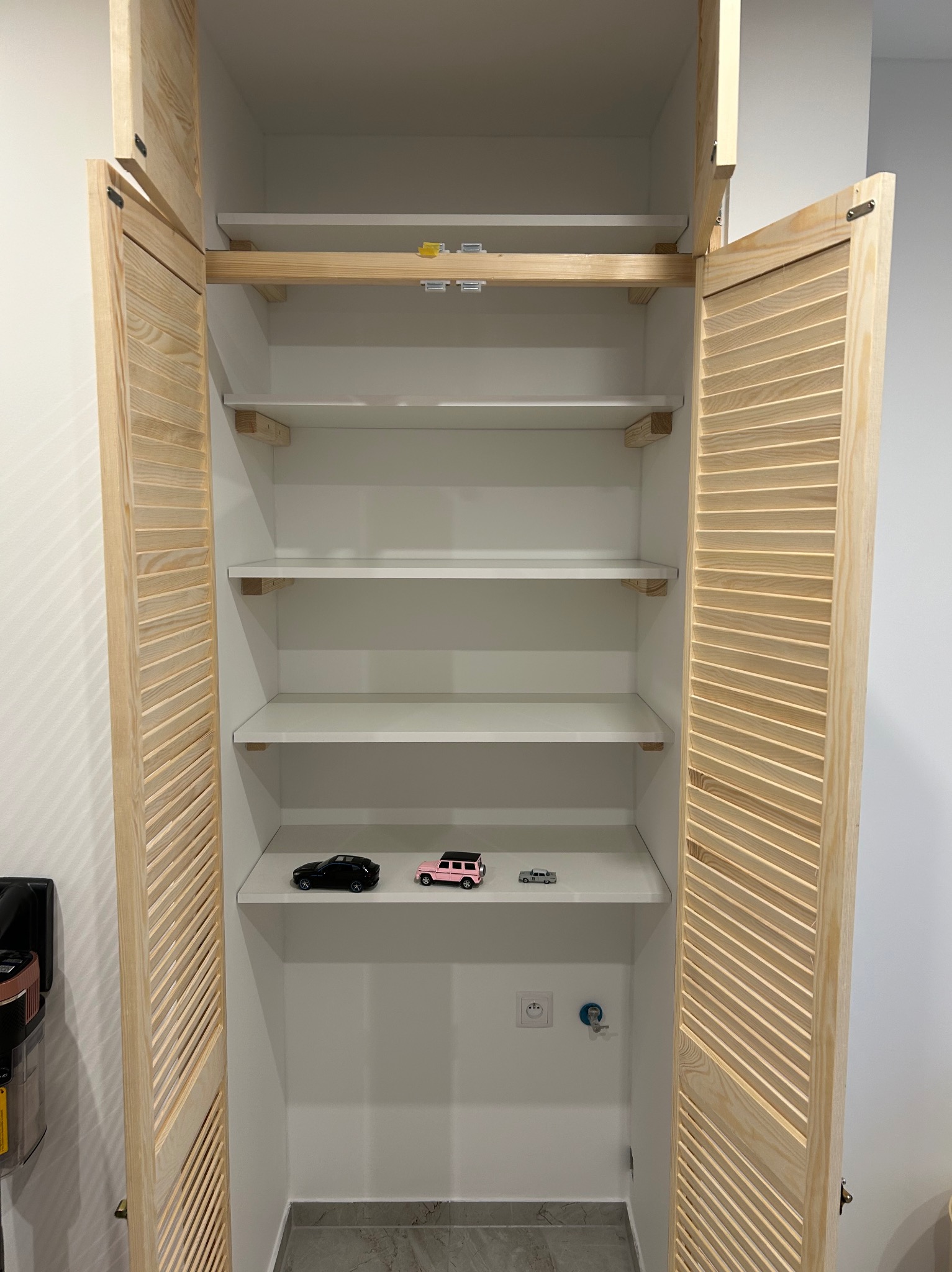 Wnętrze białej szafy z drewnianymi, żaluzjowymi drzwiami, na półce trzy zabawki - modele samochodów: czarny, różowy i srebrny.