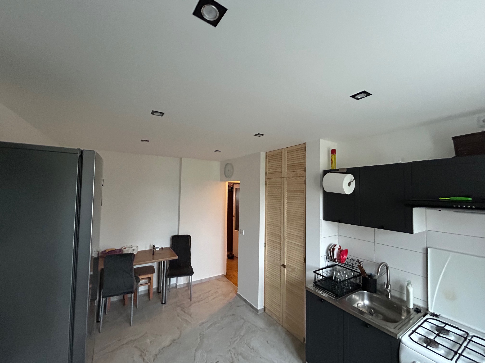 Nowoczesna kuchnia z czarnymi szafkami i białymi płytkami, widoczny stół jadalny z krzesłami w tle, marmurowa podłoga i oświetlenie sufitowe.