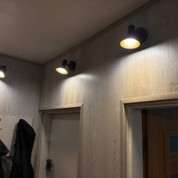 ElektroPakiet - Trzy czarne lampy punktowe zamontowane na ścianie z panelami imitującymi drewno, oświetlające wejście do pomieszczenia z dwiema parami drzwi.