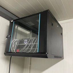 ElektroPakiet - Czarna szafa rackowa z przeszklonymi drzwiami, zamontowana na ścianie z białych paneli, widoczne białe kable wewnątrz szafy, zamek przy drzwiach.