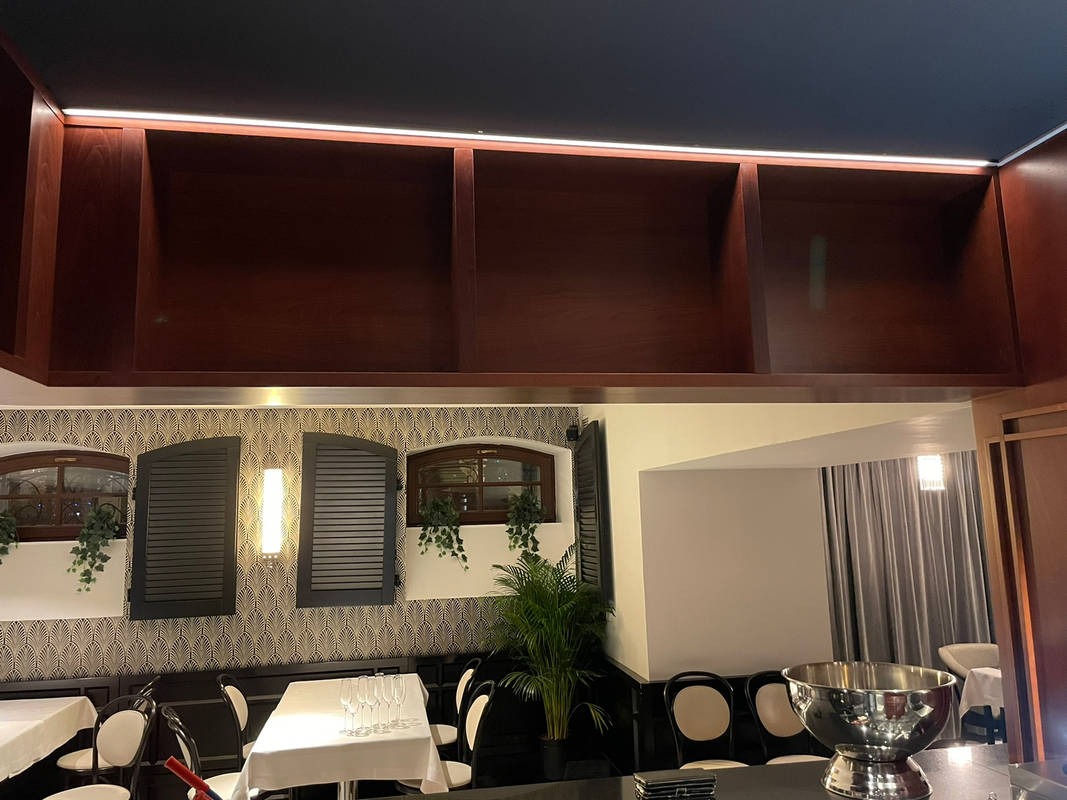 Wnętrze eleganckiej restauracji z drewnianymi szafkami pod sufitem, oświetleniem LED, oknami z okiennicami i dekoracyjną tapetą w stylu art deco.