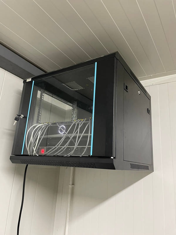 Czarna szafa rackowa z przeszklonymi drzwiami, zamontowana na ścianie z białych paneli, widoczne białe kable wewnątrz szafy, zamek przy drzwiach.