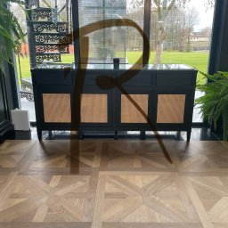 Sztuka Tworzenia Mebli - Czarna komoda z rattanowymi frontami, ustawiona na parkiecie w jodełkę, widok na ogród przez duże okno, dekorowana roślinami doniczkowymi i spiralnymi schodami.