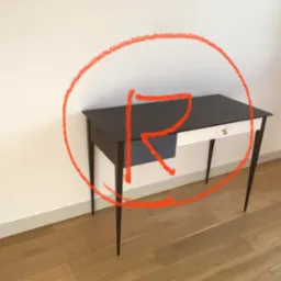 Minimalistyczny stolik z ciemnym blatem, jasnym frontem szuflady i smukłymi nóżkami, stojący na drewnianej podłodze przy białej ścianie, oznaczony okręgiem z czerwoną literą R.