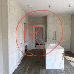 Nowoczesna kuchnia z białymi, klasycznymi frontami szafek, marmurowym blatem wyspy i złotą armaturą, widok na sąsiednie pomieszczenie z obuwiem.