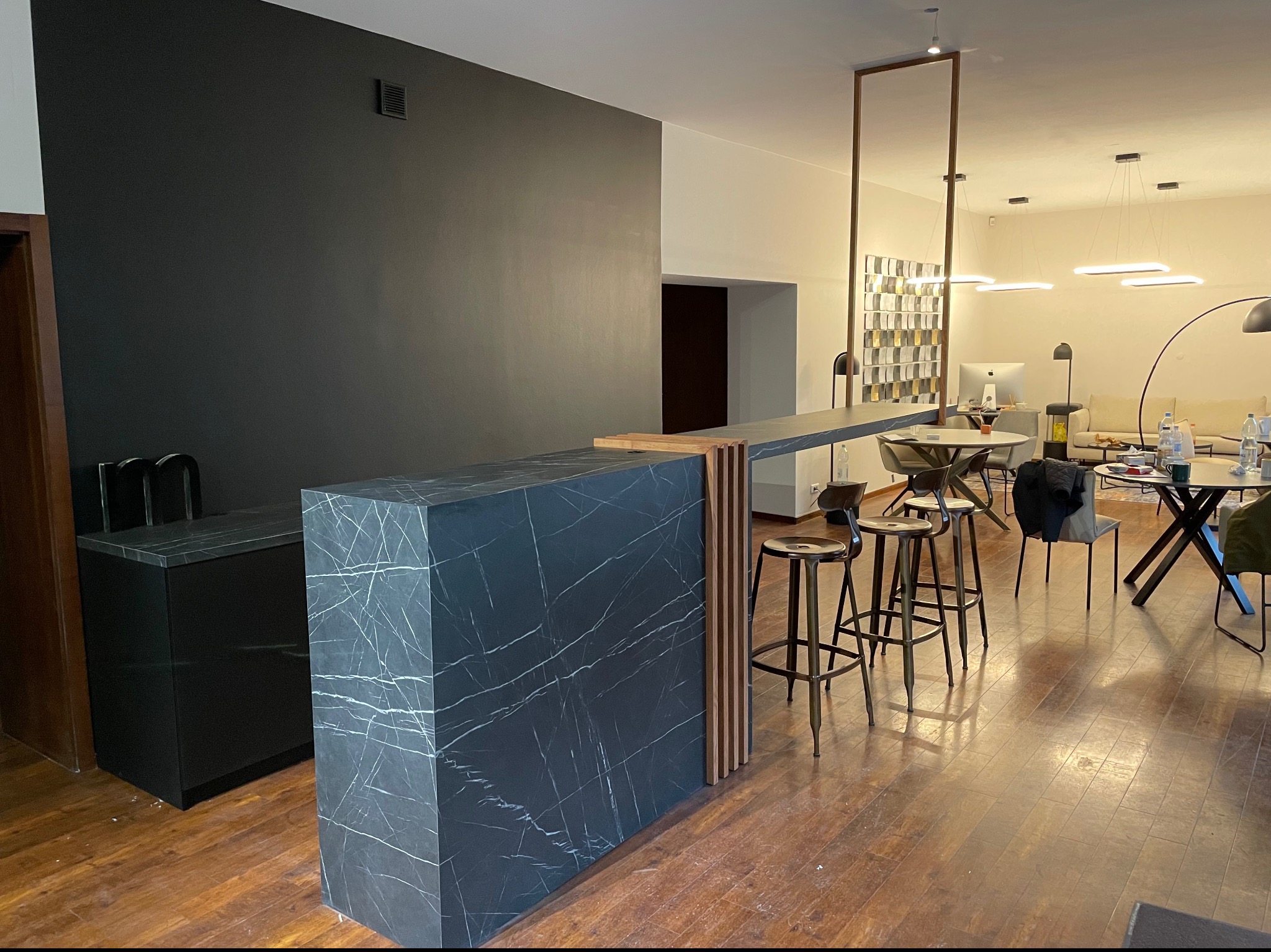 Nowoczesna wyspa kuchenna z czarnego marmuru z białymi żyłami, drewniane lamele dekoracyjne, wysokie stołki barowe w tle, widok na salon z oświetleniem.