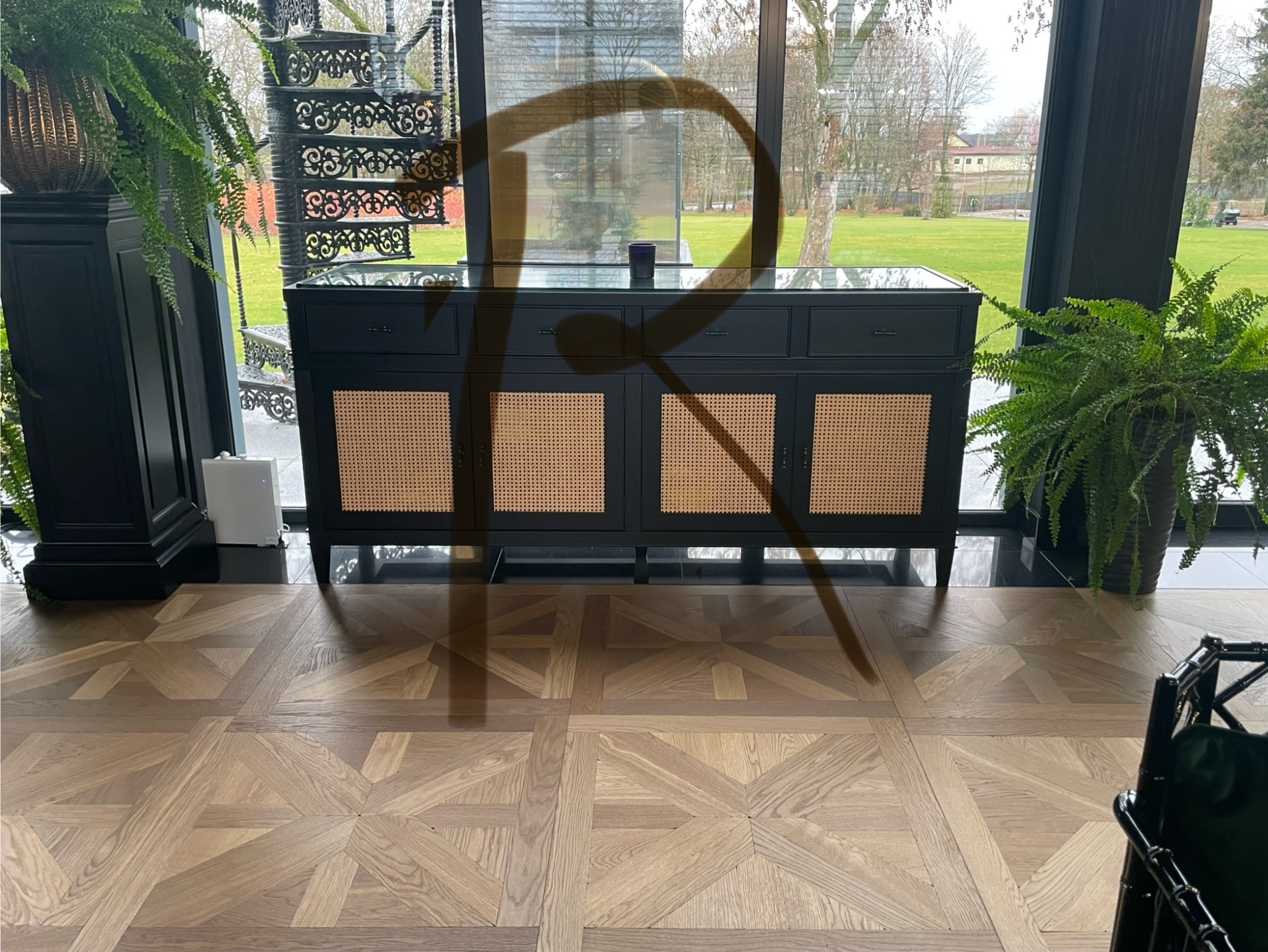 Czarna komoda z rattanowymi frontami, ustawiona na parkiecie w jodełkę, widok na ogród przez duże okno, dekorowana roślinami doniczkowymi i spiralnymi schodami.