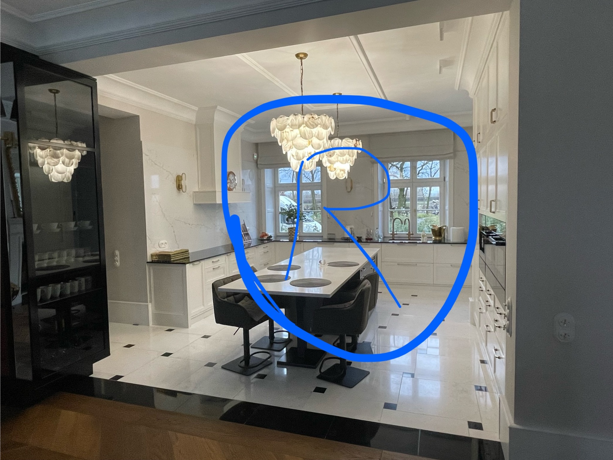 Elegancka, jasna kuchnia z wyspą, oświetlona dwoma dekoracyjnymi żyrandolami; widoczna zabudowa meblowa z białymi frontami, marmurowy blat i podłoga w czarno-białą szachownicę.