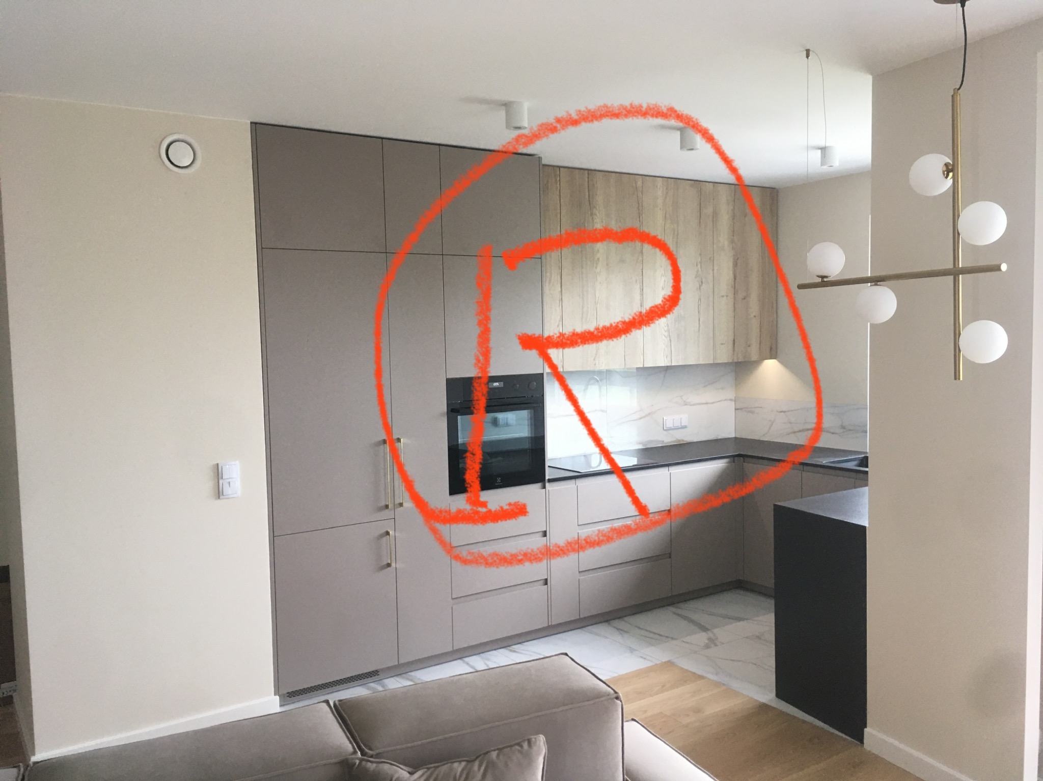 Nowoczesna kuchnia na wymiar w odcieniach beżu i drewna, z czarnym piekarnikiem i marmurowym blatem, oświetlona designerską lampą wiszącą.