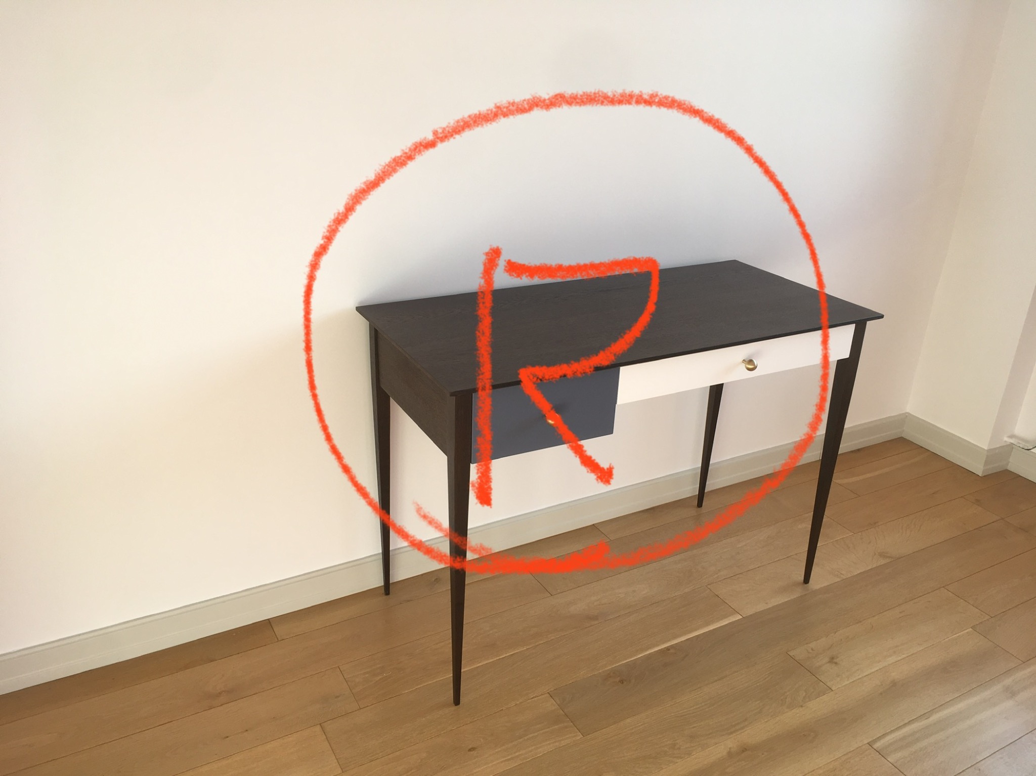 Minimalistyczny stolik z ciemnym blatem, jasnym frontem szuflady i smukłymi nóżkami, stojący na drewnianej podłodze przy białej ścianie, oznaczony okręgiem z czerwoną literą R.