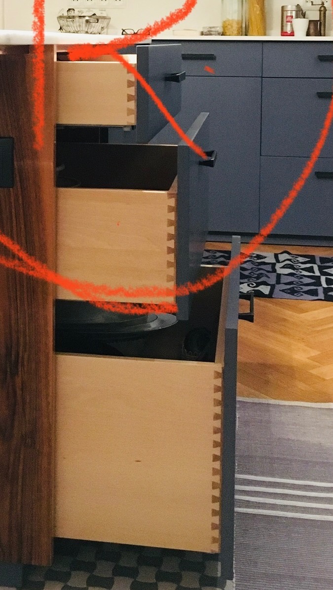 Otwarta szuflada z widocznymi połączeniami stolarskimi typu jaskółczy ogon, w tle zabudowa kuchenna w kolorze granatowym, widoczny fragment podłogi z drewna i dywanu.