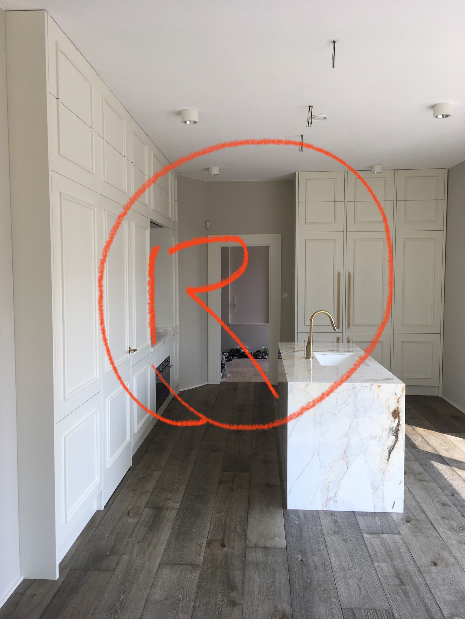 Nowoczesna kuchnia z białymi, klasycznymi frontami szafek, marmurowym blatem wyspy i złotą armaturą, widok na sąsiednie pomieszczenie z obuwiem.