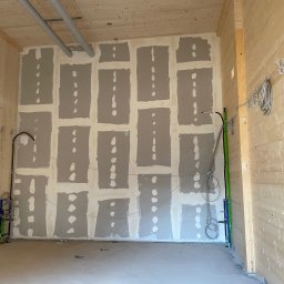 Kacper Siwiec - Wnętrze pomieszczenia w trakcie prac wykończeniowych: ściana z płyt kartonowo-gipsowych z widocznymi łączeniami szpachlowanymi punktowo, drewniane panele ścienne, instalacje elektryczne i rury...