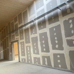 Kacper Siwiec - Wysoka: Ściana w trakcie przygotowania do malowania, z widocznymi łączeniami płyt gipsowo-kartonowych i metalowymi korytkami kablowymi, drewniana konstrukcja sufitu i ścian w tle, dwóch pracowników...