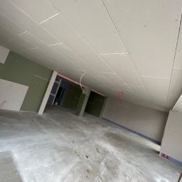 Kacper Siwiec - Montaż płyt kartonowo-gipsowych na suficie i ścianach w surowym stanie, widoczne kable wystające z sufitu, betonowa podłoga z zabrudzeniami, perspektywa z dołu.