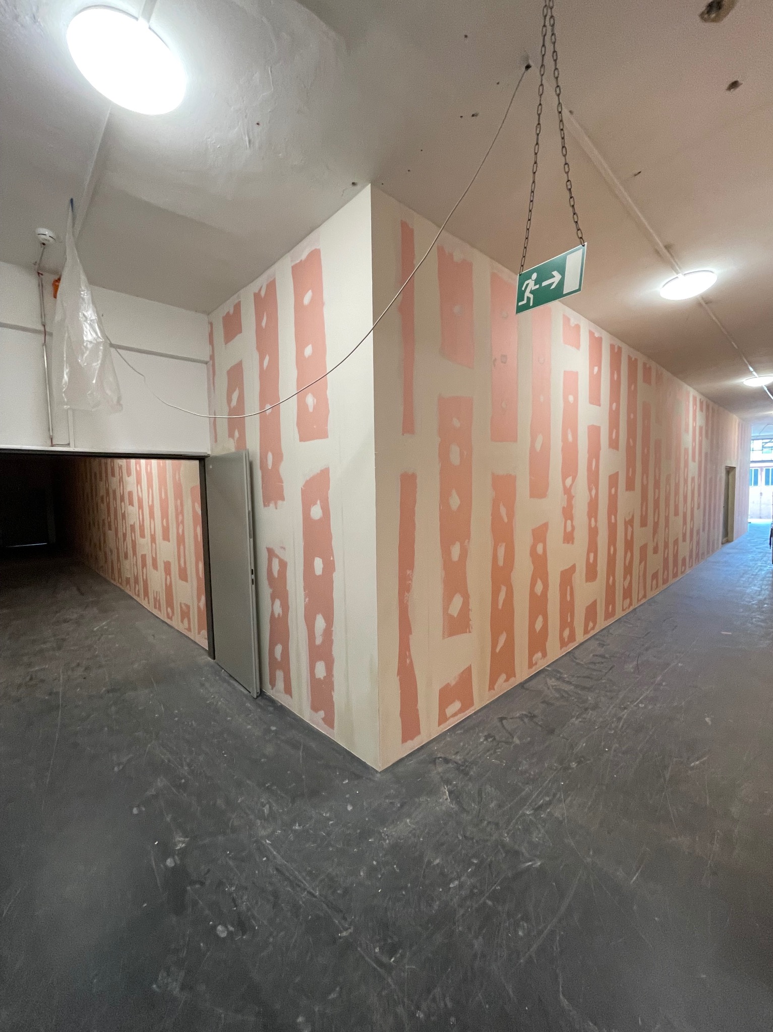 Korytarz w budynku komercyjnym z przygotowanymi ścianami do dalszych prac wykończeniowych, widoczne łączenia płyt gipsowych pokryte masą szpachlową, znak ewakuacyjny podwieszony na łańcuchu.