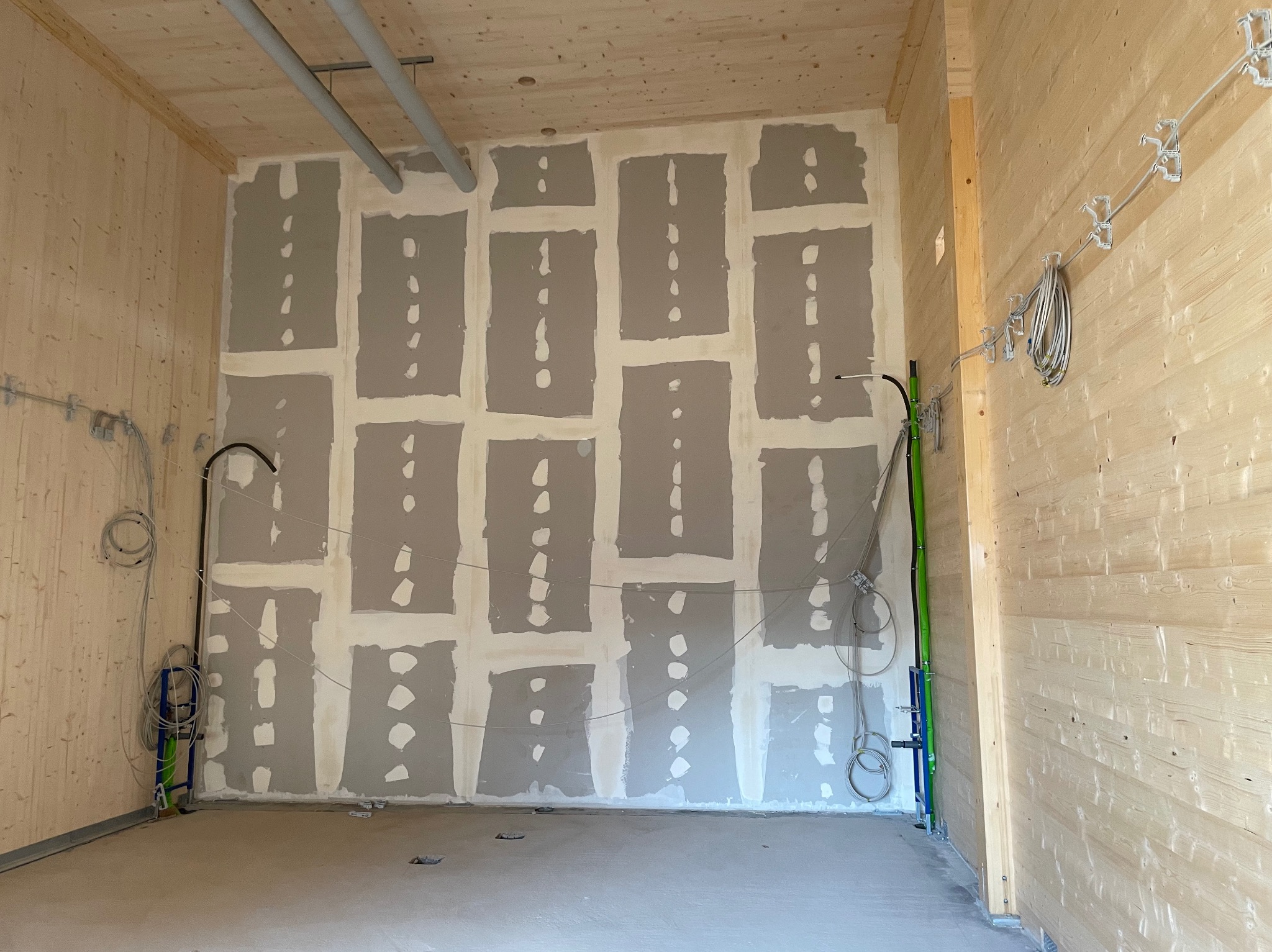 Wnętrze pomieszczenia w trakcie prac wykończeniowych: ściana z płyt kartonowo-gipsowych z widocznymi łączeniami szpachlowanymi punktowo, drewniane panele ścienne, instalacje elektryczne i rury...