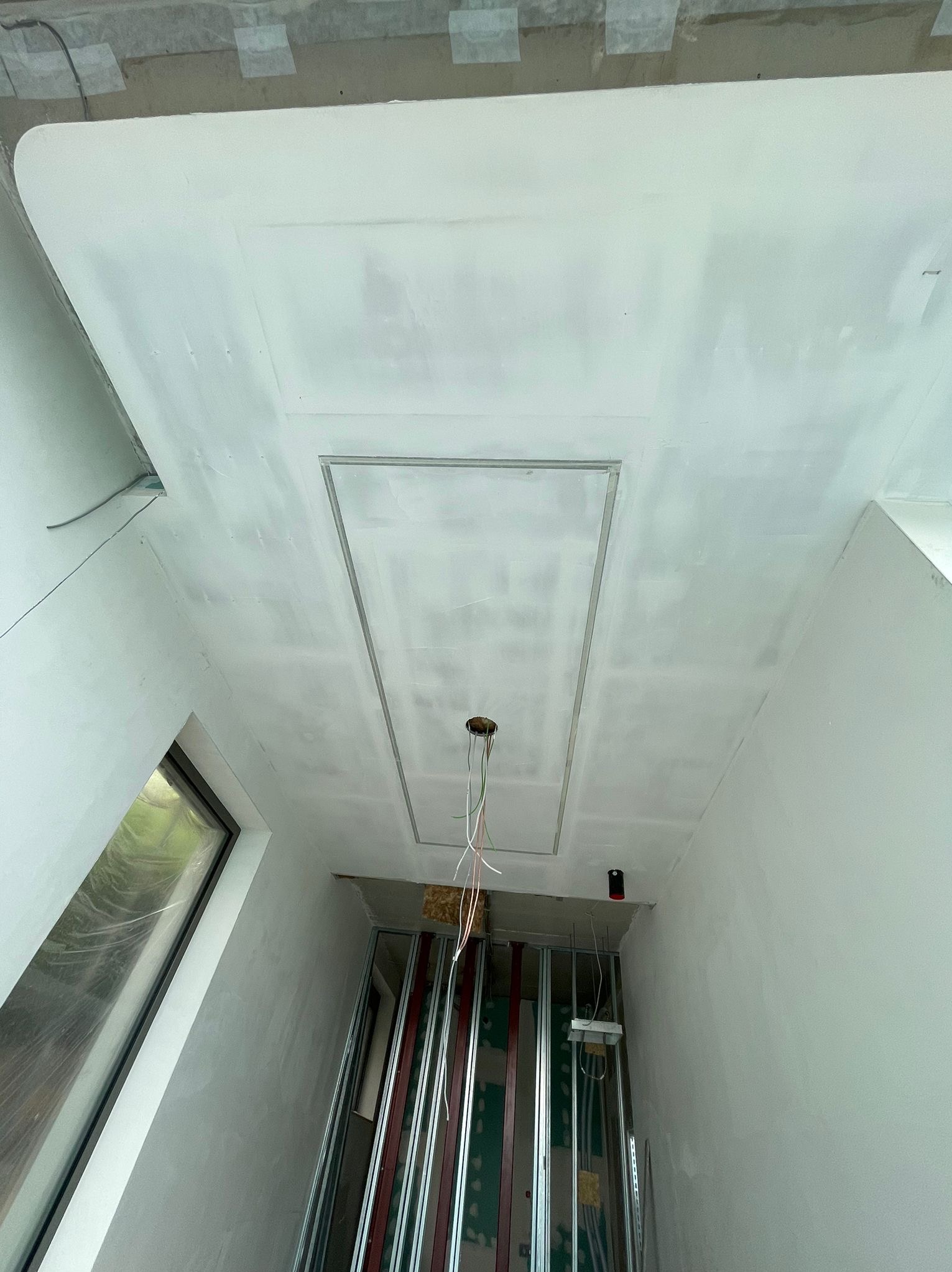 Sufit z wykończoną gładzią, z widocznym otworem rewizyjnym obramowanym metalową listwą oraz wystającymi przewodami elektrycznymi, poniżej szkielet konstrukcji ścianki działowej z profili metalowych.