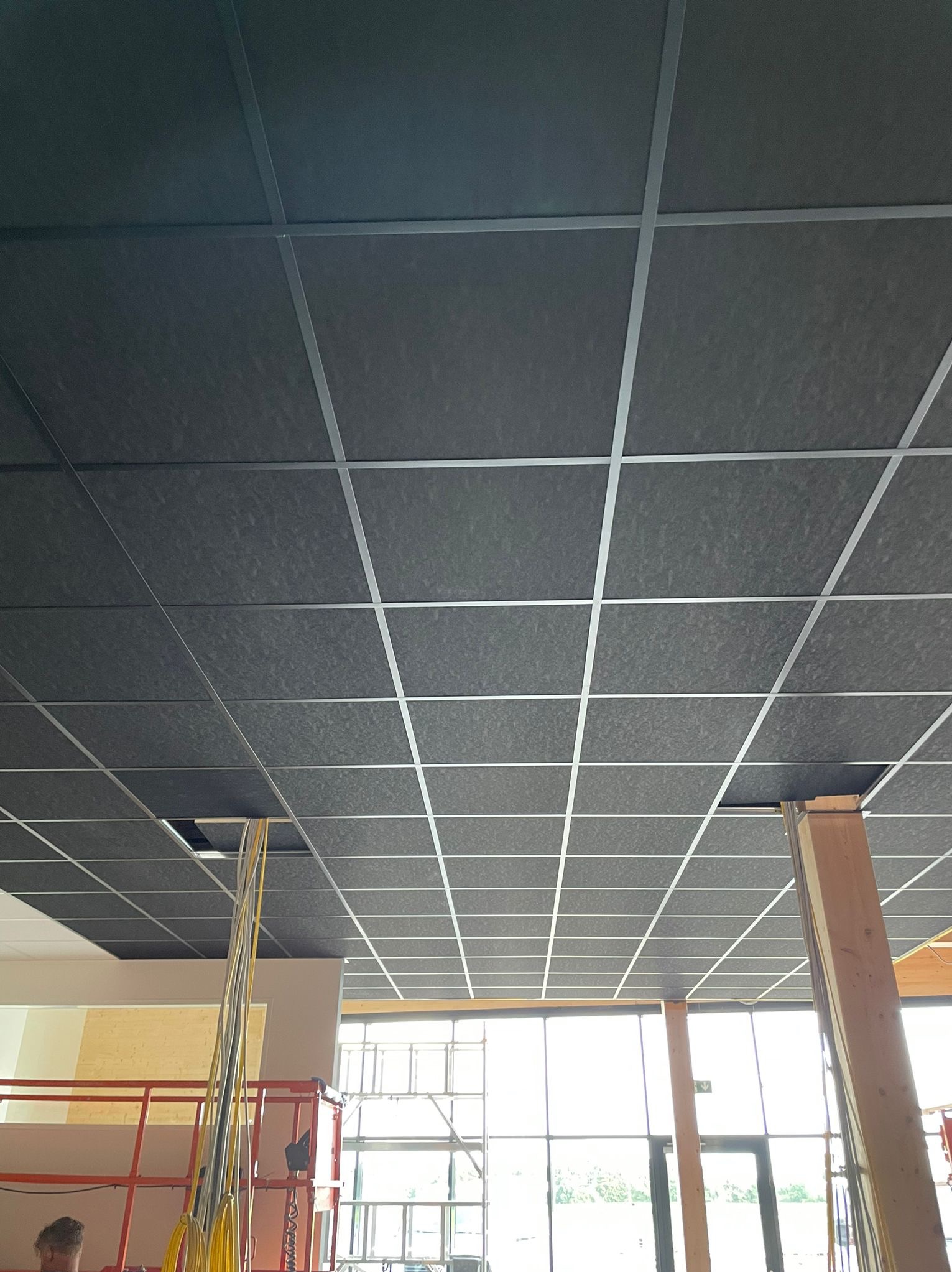 Montaż sufitu podwieszanego z czarnych paneli w rastrze z metalowych profili, widoczne przewody wystające z otworu, fragment rusztowania i okna w tle.