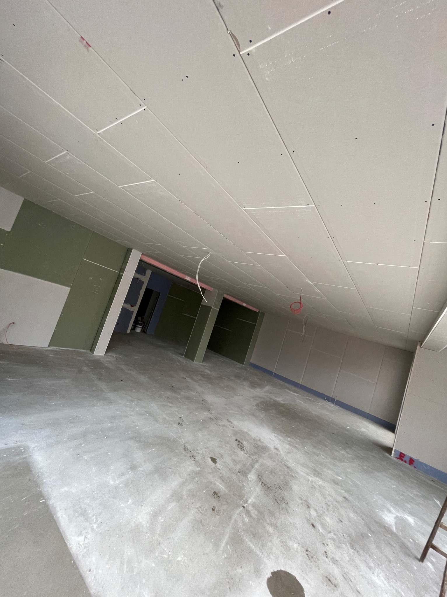 Montaż płyt kartonowo-gipsowych na suficie i ścianach w surowym stanie, widoczne kable wystające z sufitu, betonowa podłoga z zabrudzeniami, perspektywa z dołu.