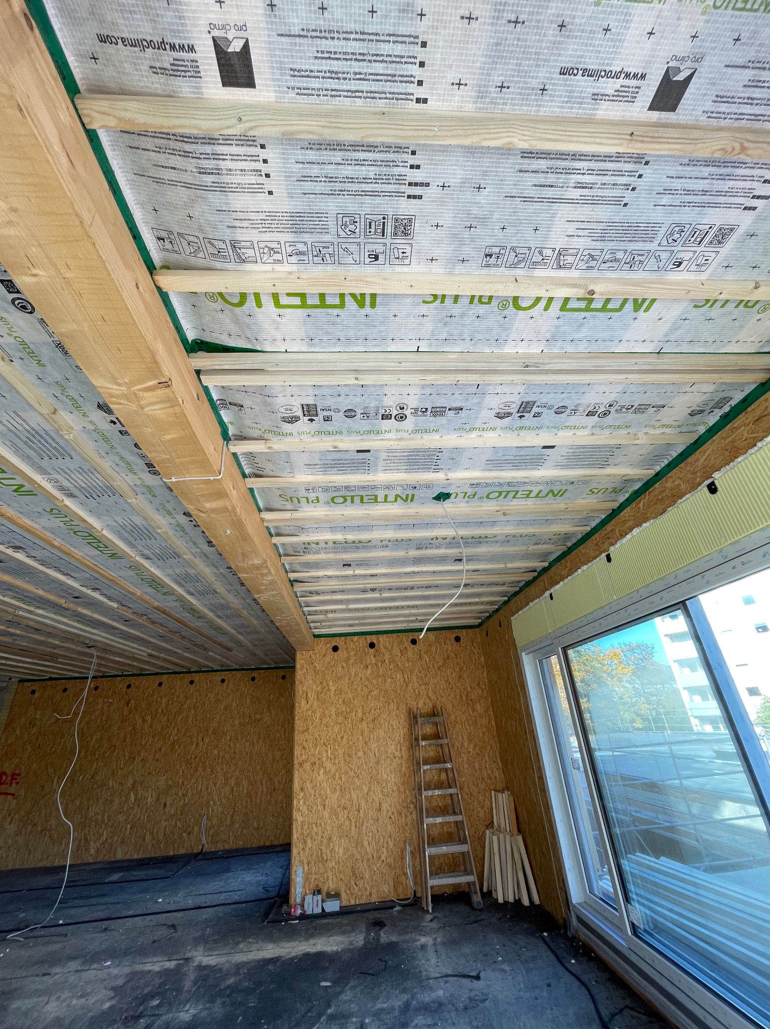 Poddasze w trakcie ocieplania, widoczne drewniane belki stropowe z folią paroizolacyjną Intelio Plus, ściany wykończone płytami OSB, drabina oparta o ścianę, okno z widokiem na budynek.