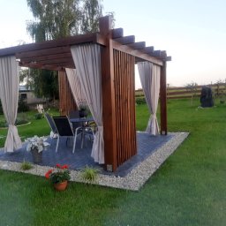 Magiczne Ogrody Kamil Gigielewicz - Drewniana altana z zasłonami na brukowanym tarasie otoczonym żwirem, z widokiem na zadbany trawnik i elementy ogrodu.