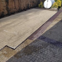 Magiczne Ogrody Kamil Gigielewicz - Nierównomiernie ułożona trawa w rolkach na przygotowanym podłożu, obok widoczna kostka brukowa i zbiornik na gaz w ogrodzie z drewnianym płotem w tle.