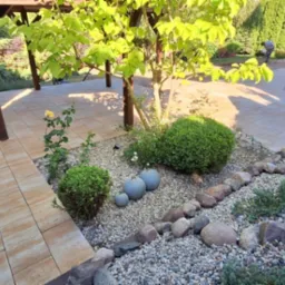 Ogród z jasną kostką brukową, rabatami z kamyków i zielenią. W tle pergola i pojazd dostawczy. Ujęcie z perspektywy ogrodu, w słoneczny dzień.