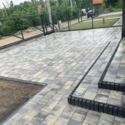 Ułożona kostka brukowa w odcieniach szarości prowadząca do domu, z widocznymi schodami i fragmentem ogrodzenia. Przygotowane miejsce pod trawnik.
