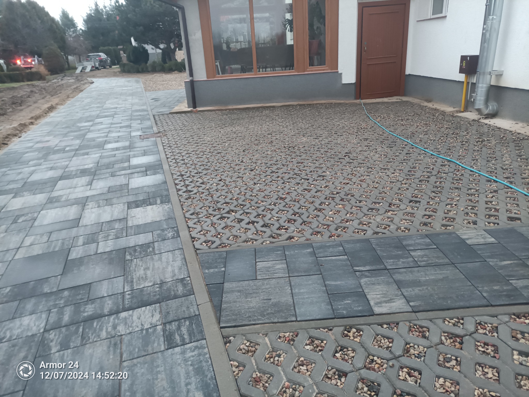 Ułożona nawierzchnia z szarej kostki brukowej o różnych wzorach, w tym ażurowej z wypełnieniem żwirowym, prowadząca do budynku z brązowymi drzwiami i dużym oknem. Widoczny fragment węża ogrodowego.