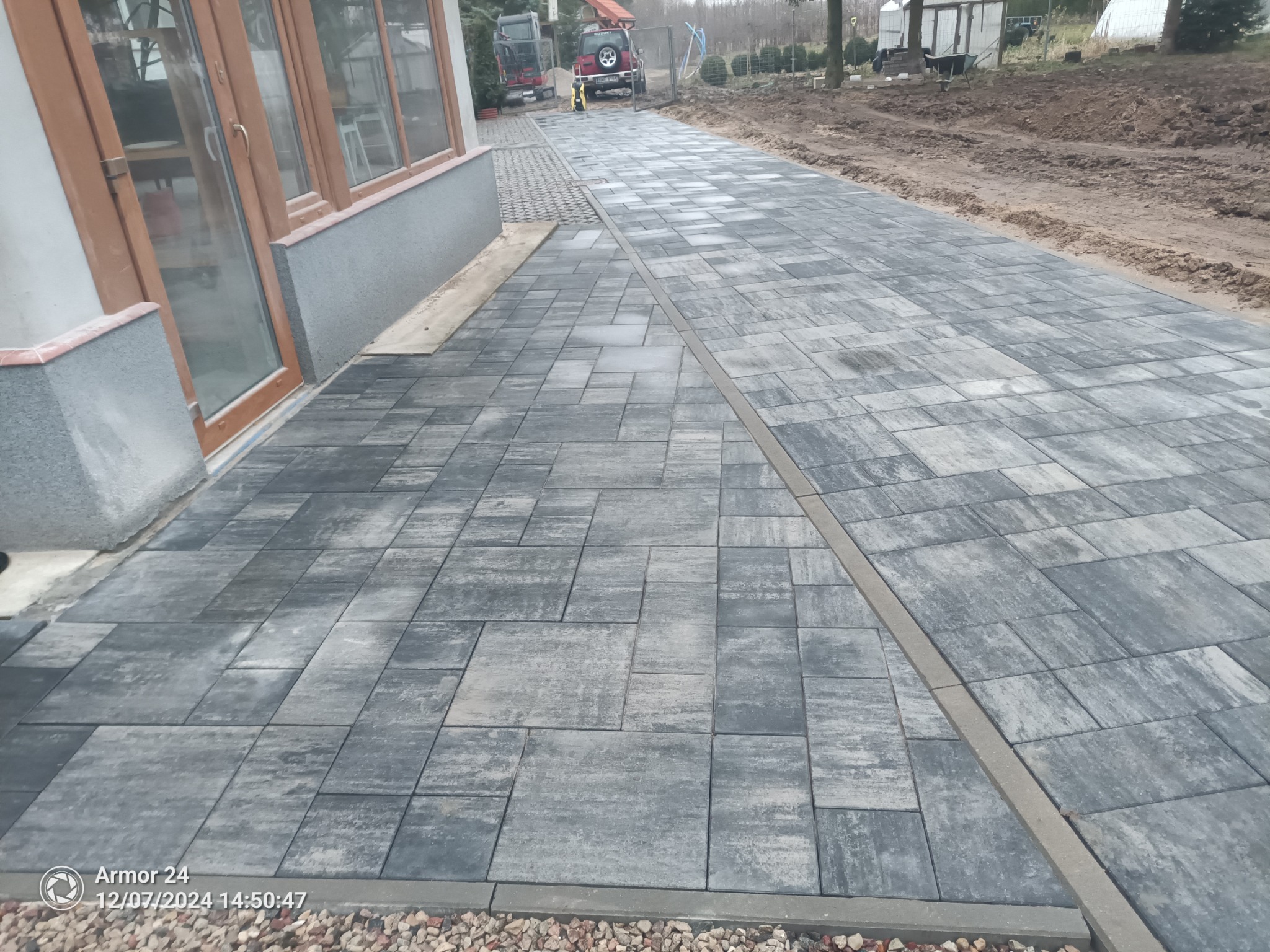 Ułożona szara kostka brukowa prowadząca od domu do drogi, z widocznymi maszynami budowlanymi w tle i granicą z kamieni ozdobnych.