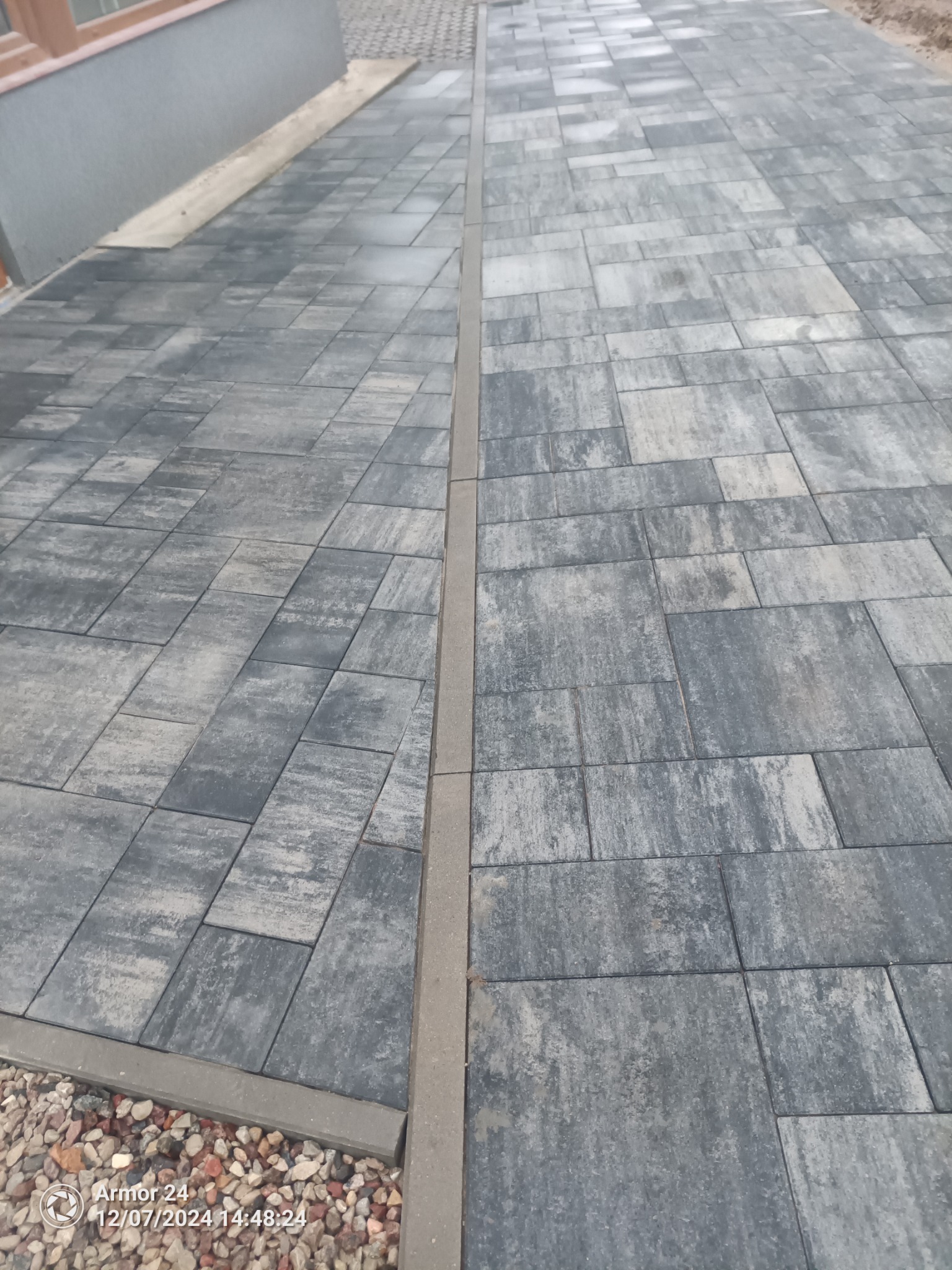 Ułożona nawierzchnia z szarej kostki brukowej o zróżnicowanych kształtach, oddzielona liniowym odwodnieniem, z widocznymi drobnymi kamieniami na krawędzi.