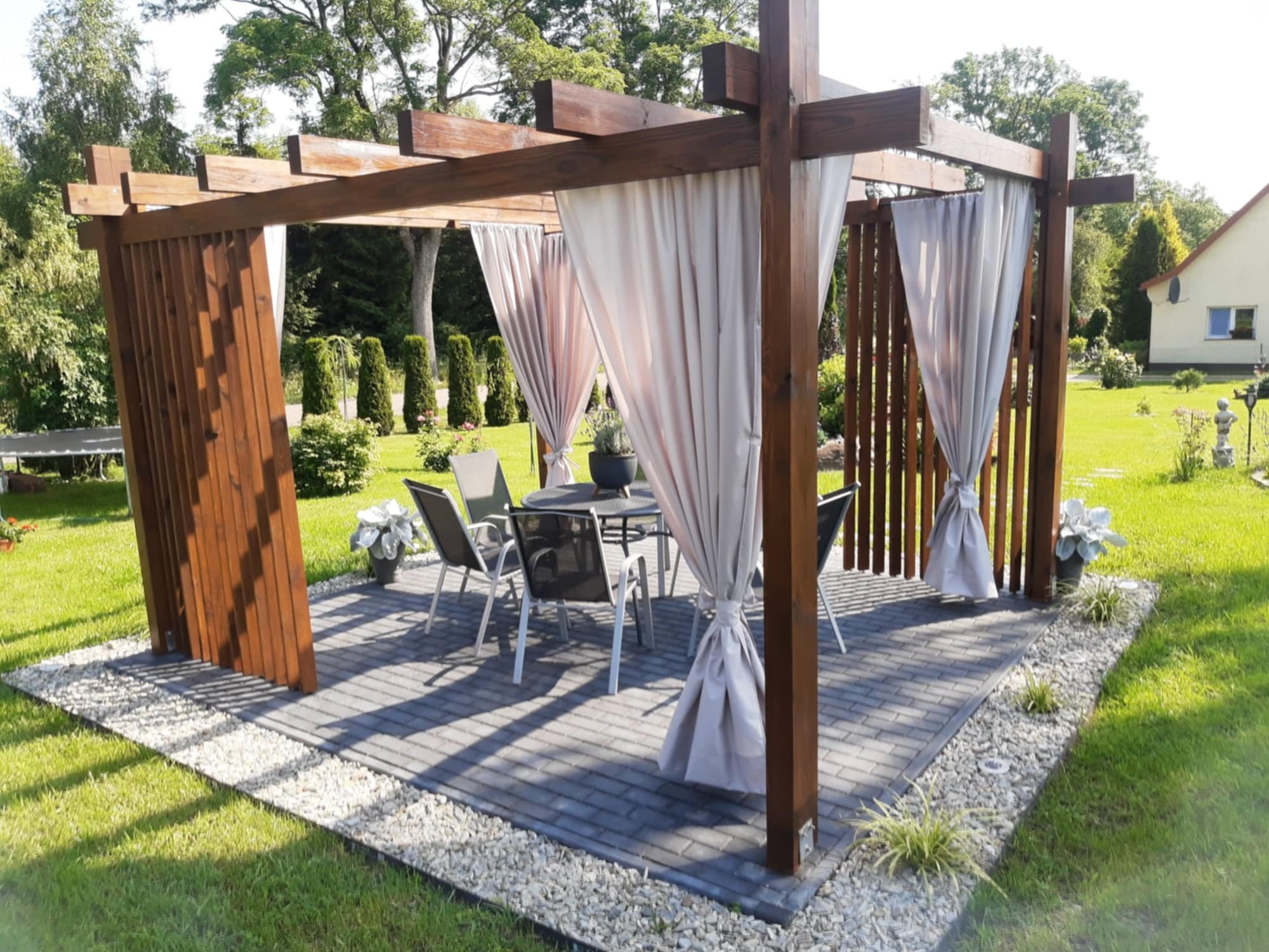Drewniana pergola z zasłonami na tarasie z szarej kostki brukowej, otoczona białym żwirem i trawnikiem, w tle dom i drzewa.