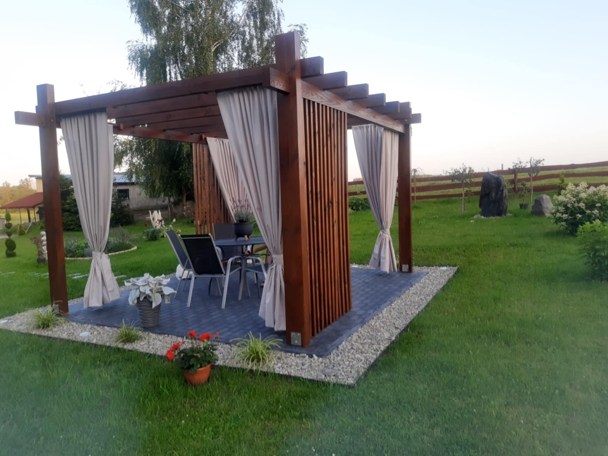 Drewniana altana z zasłonami na brukowanym tarasie otoczonym żwirem, z widokiem na zadbany trawnik i elementy ogrodu.