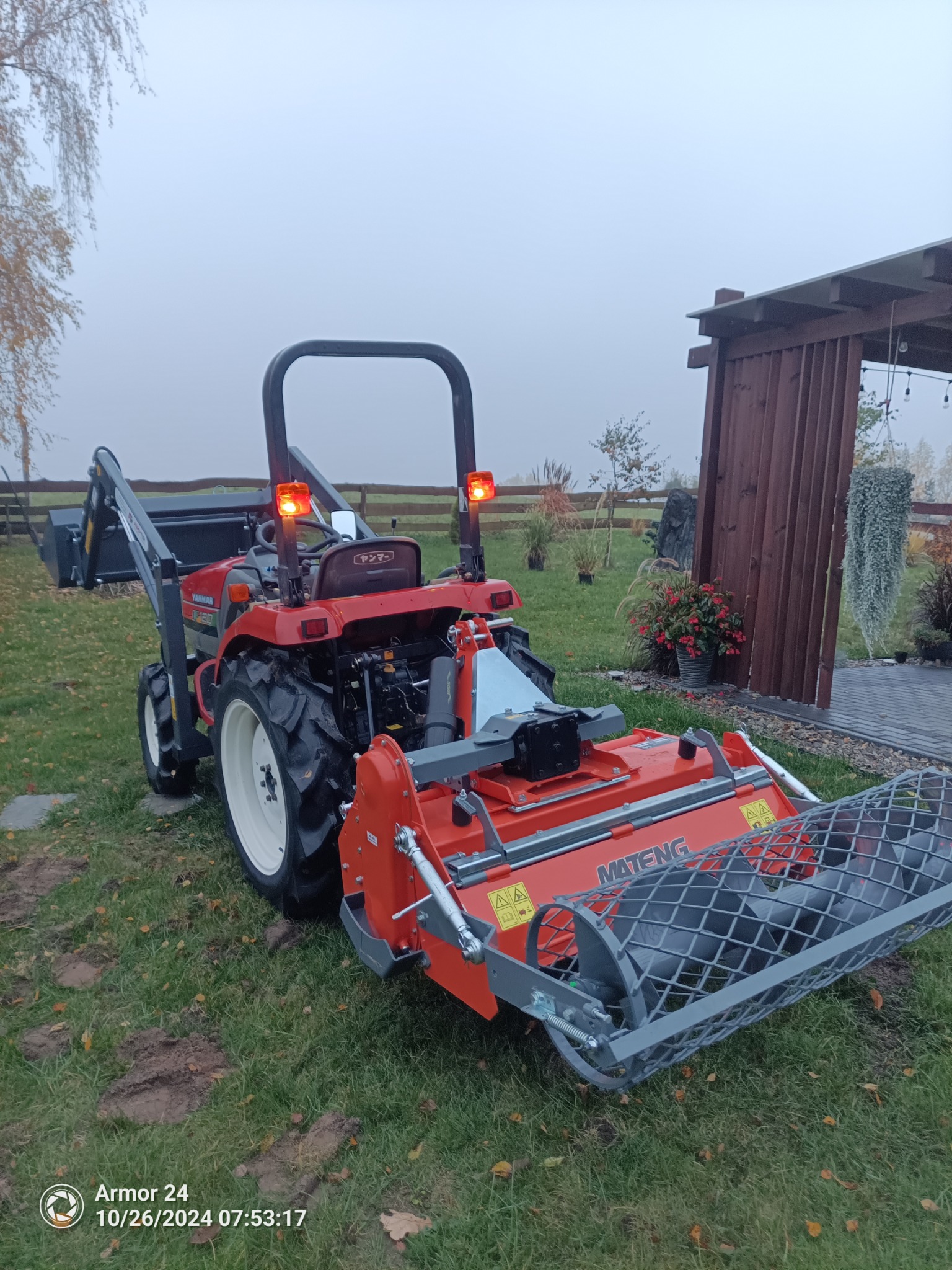 Czerwony traktor Yanmar z glebogryzarką Mateng na trawniku z widocznymi śladami opon, w tle drewniana pergola i rośliny ozdobne.