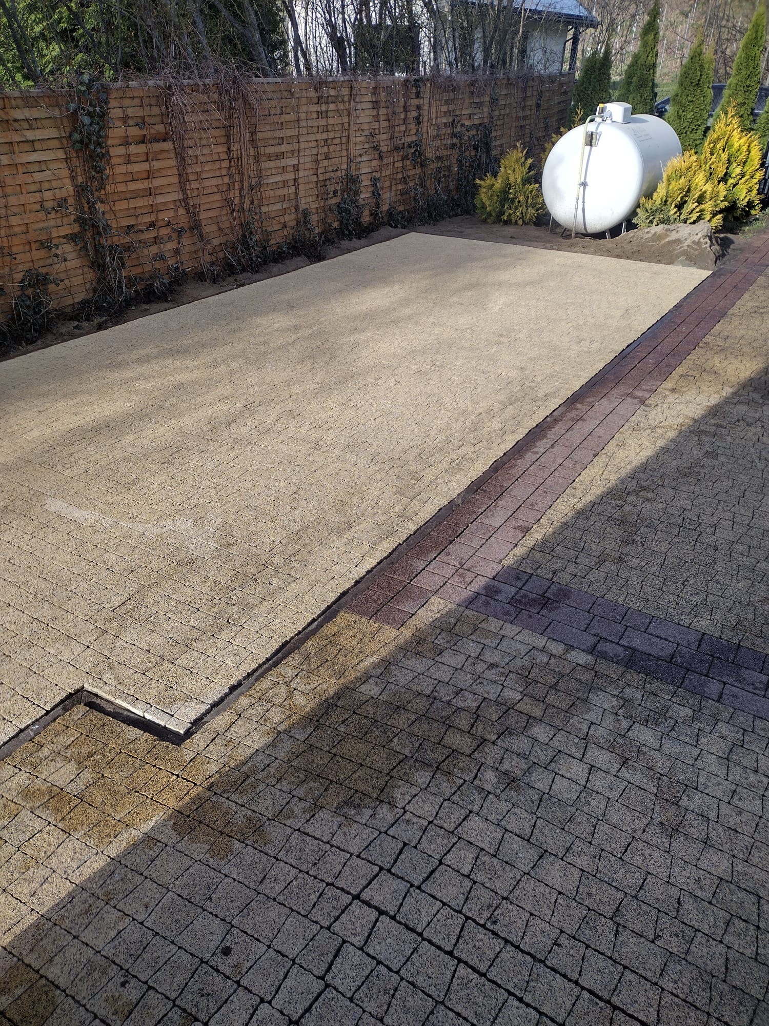 Nierównomiernie ułożona trawa w rolkach na przygotowanym podłożu, obok widoczna kostka brukowa i zbiornik na gaz w ogrodzie z drewnianym płotem w tle.