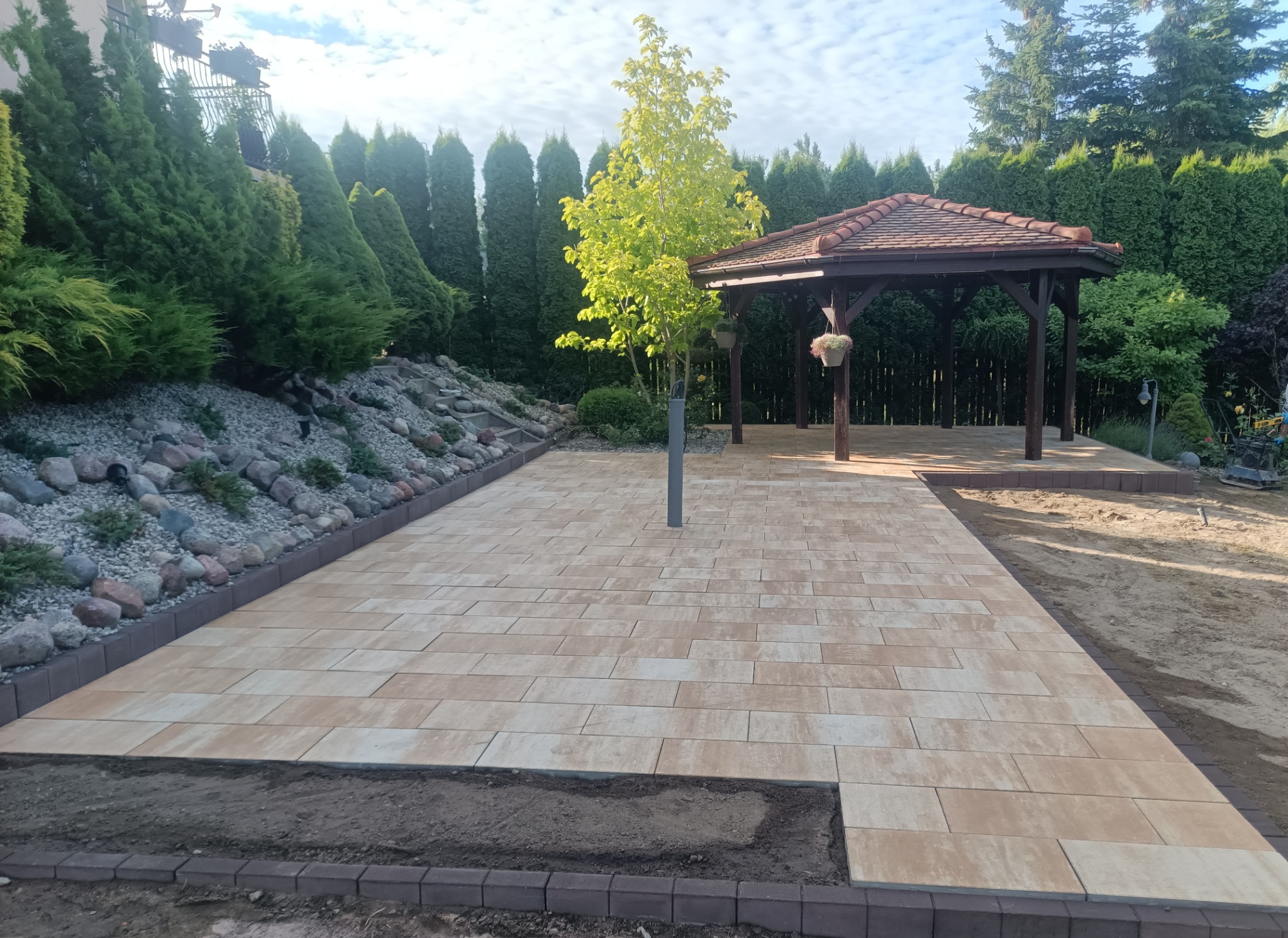 Jasna kostka brukowa ułożona wokół drzewa i altany, z obrzeżami z ciemnej kostki. W tle iglaki i kamienie. Częściowo widoczna niezakończona praca.