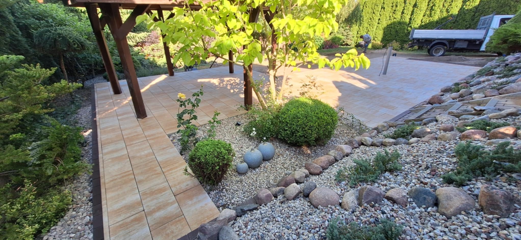 Ogród z jasną kostką brukową, rabatami z kamyków i zielenią. W tle pergola i pojazd dostawczy. Ujęcie z perspektywy ogrodu, w słoneczny dzień.