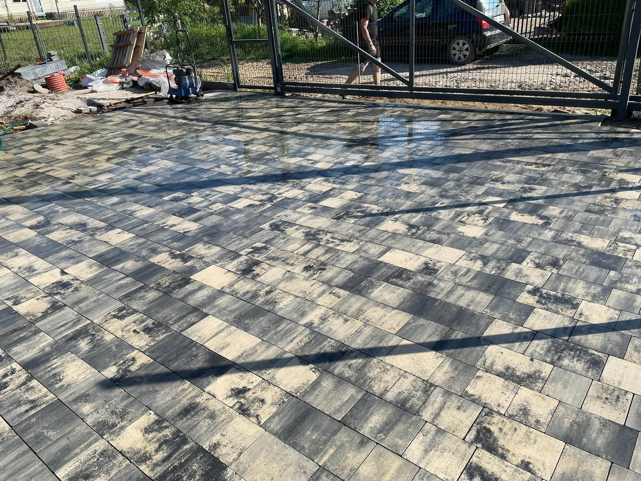 Ułożona kostka brukowa w odcieniach szarości i beżu, z widocznym fragmentem metalowej bramy w tle i ubijarką w lewym górnym rogu. Częściowo mokra nawierzchnia.