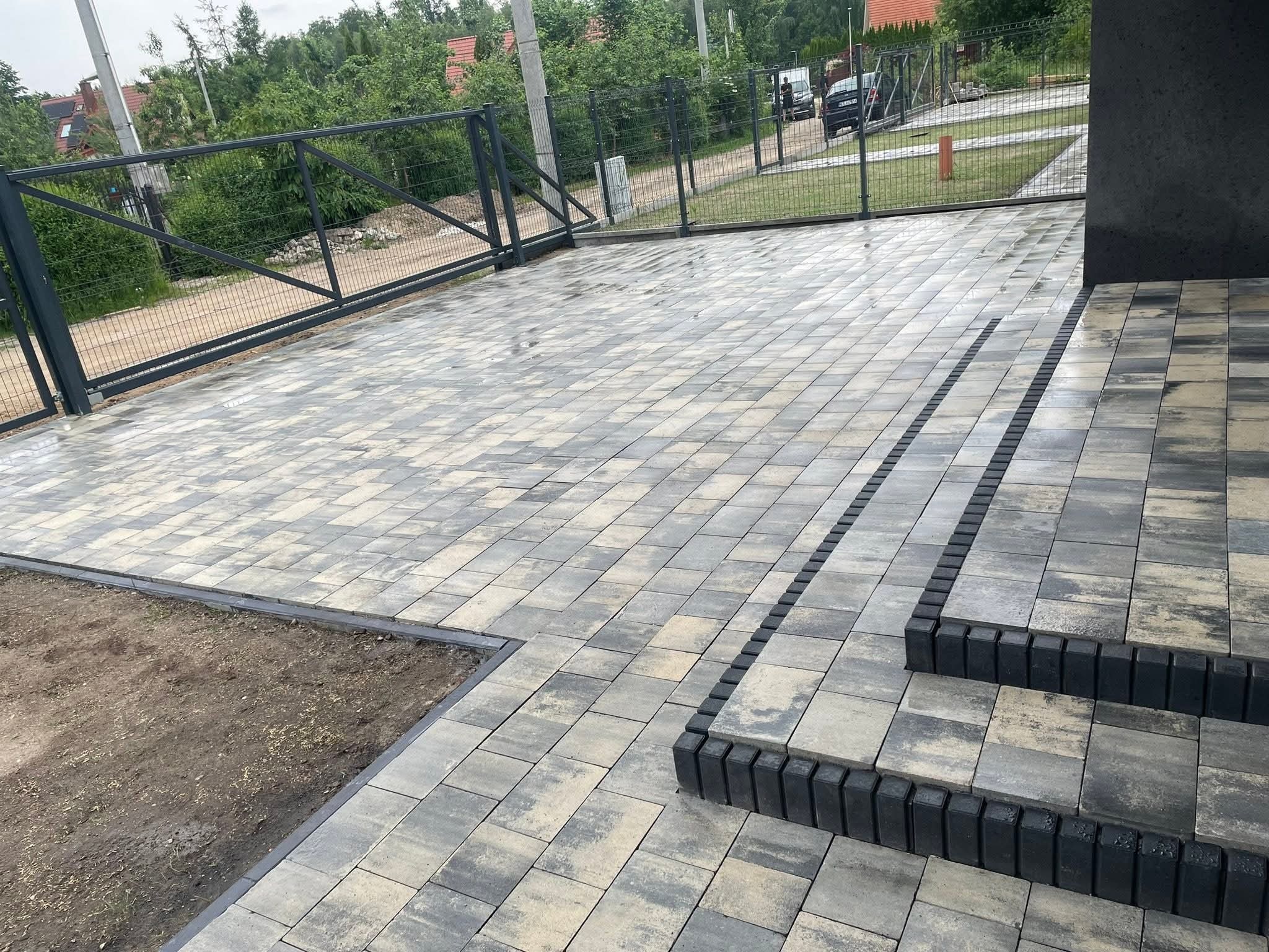 Ułożona kostka brukowa w odcieniach szarości prowadząca do domu, z widocznymi schodami i fragmentem ogrodzenia. Przygotowane miejsce pod trawnik.